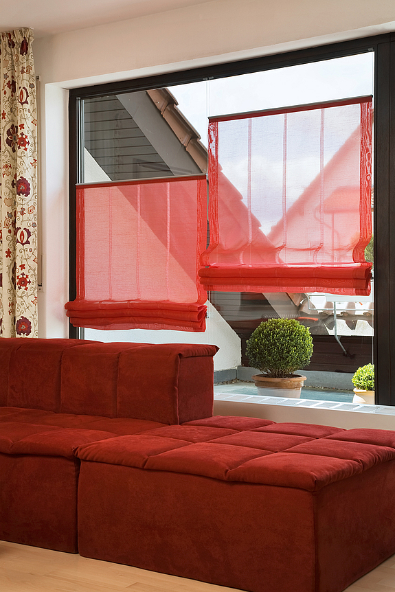 rote, halb transparente Raffrollos an einem Fenster neben einer roten Couch.