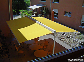 Ansicht von oben; gelbe, freistehende Markise an roter Hauswand über Tischen und Stühlen.
