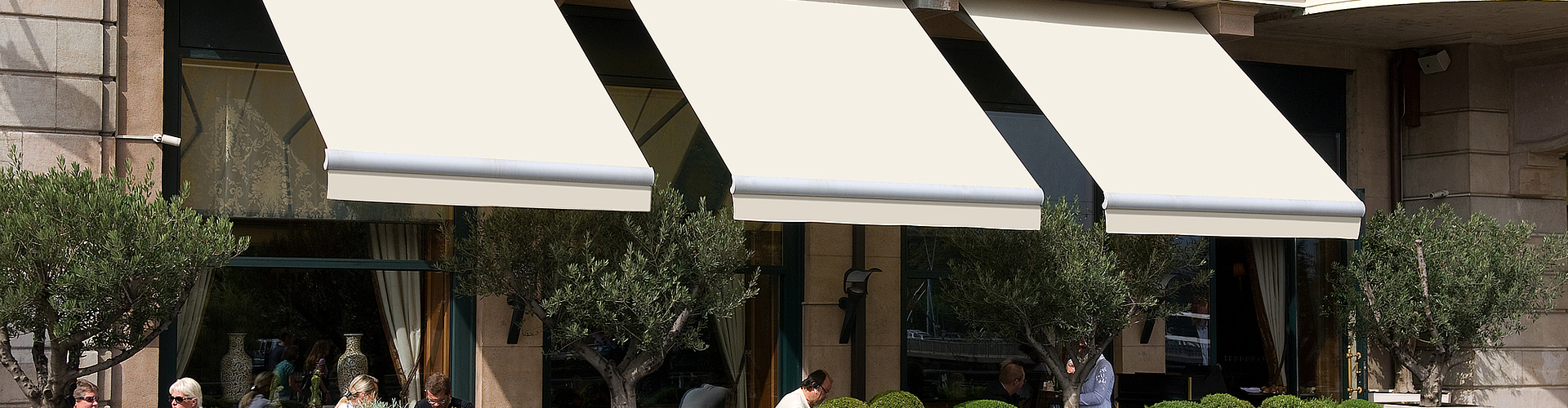 drei weiße, ausgefahrene Gelenkarm-Markise mit mehr Ausladung als Breite nebeneinander über einem Café an einem Altbau mit einigen Pflanzen.