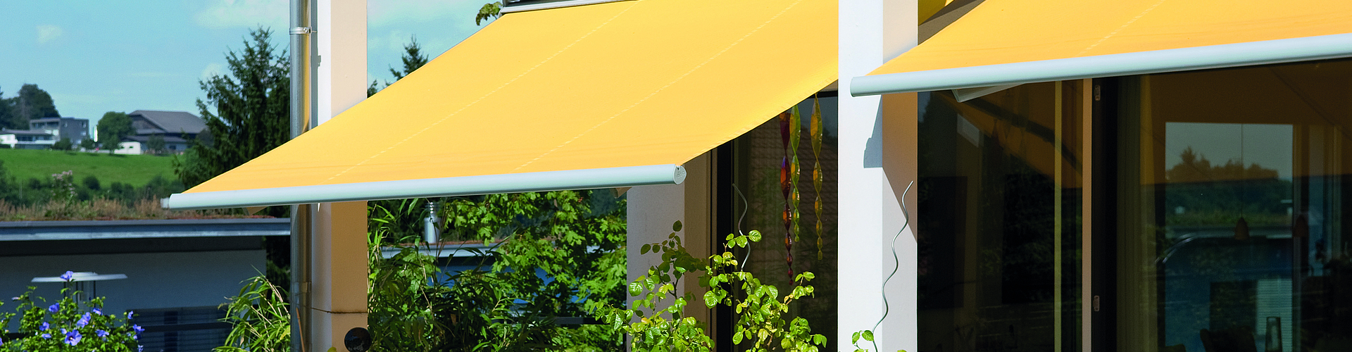 gelbe, ausgefahrene Gelenkarm-Markisen über Terrasse, getrennt durch weiße Säulen, Terrasse mit Pflanzen, Tisch und Stühlen