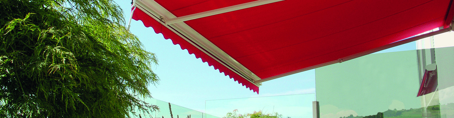 rote, ausgefahrene Gelenkarm-Markise an weißer Hauswand, mit Terrasse, Liegestühlen, Glas-Geländer und Pflanzen.