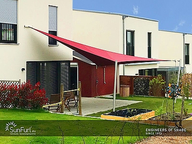 rotes, gespanntes Sunfurl-Sonnensegel, in einem kleinen, grünen Garten mit modernem, weißem Haus.